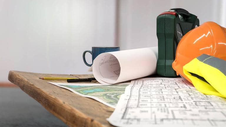 A reflective jacket, hardhat, rolled blueprint and mug placed on a wooden table