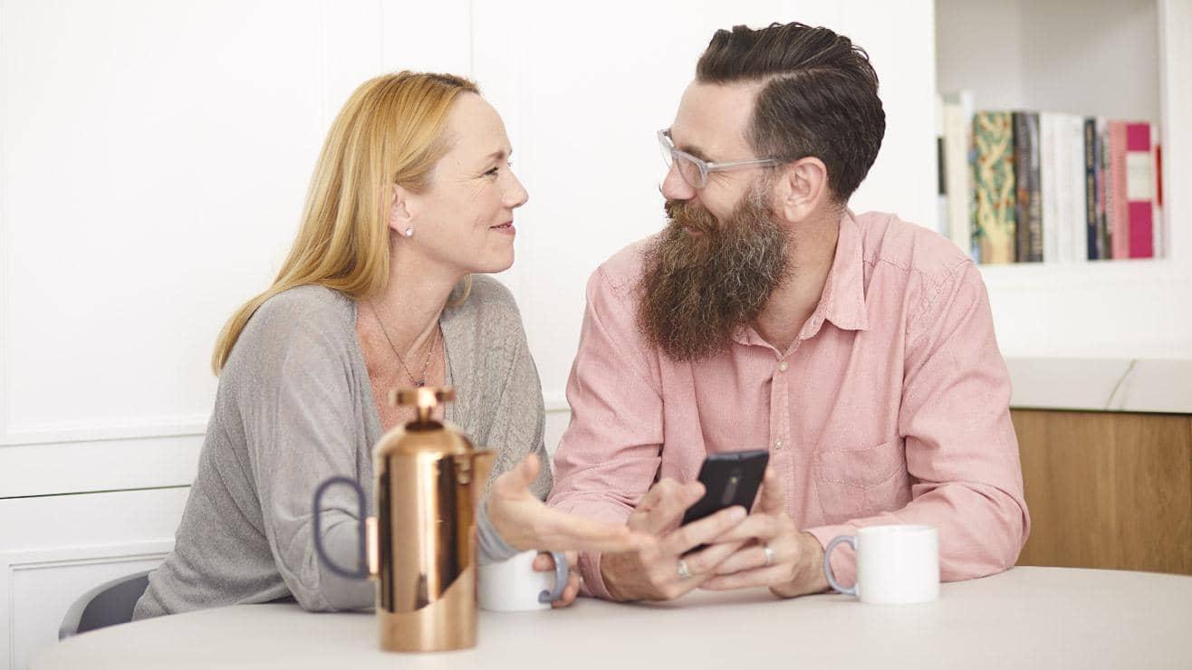 A smiling couple in a kitchen look at a mobile phone and drink coffee
