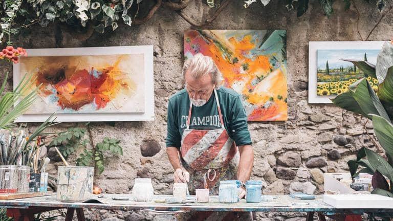  A man wearing an apron paints a canvas on a table covered in pots and brushes