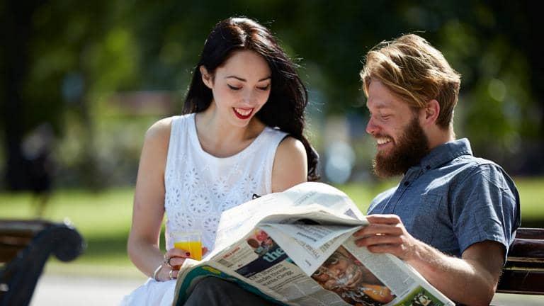 A man and a woman sitting on a park bench look at a newspaper
