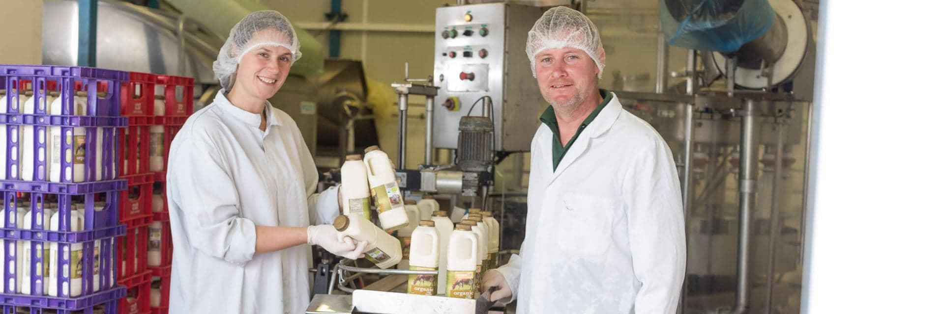  A woman and a man dressed in overalls hold up bottles of milk that have just come off the dairy production line