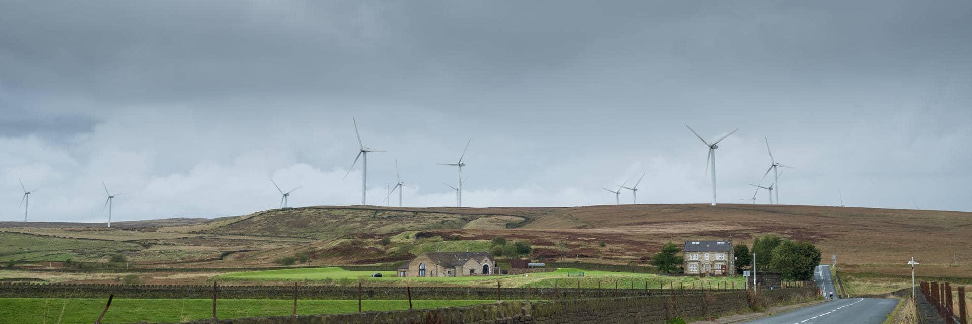 A line of wind turbines on a hillside with a road and old stone buildings in the foreground