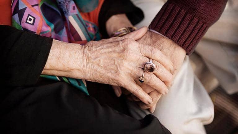  A person holds the hand of an elderly woman who is wearing rings on her fingers