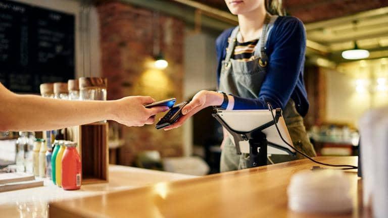 Cashier at a coffee shop takes payment from a customer using contactless payment on their mobile phone.