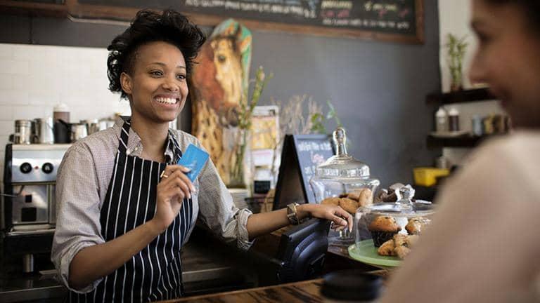 A café worker wearing an apron smiles at a customer as she holds up a bank card