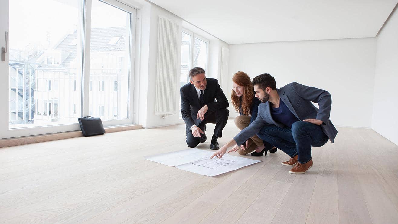 Two men and a woman dressed in professional clothes crouch down to study architectural plans laid out on the floor of an empty room
