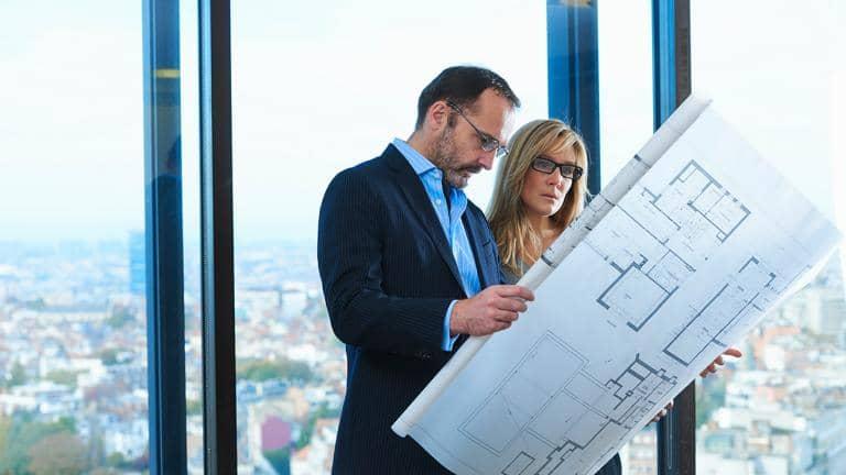 A man and a woman dressed in professional clothes study architectural plans while standing in front of a large window overlooking a town