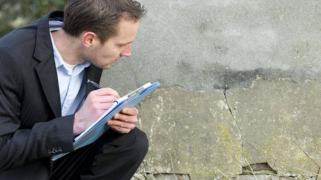 A man wearing a suit and holding a clipboard and pen crouches down to look at a crack on the outside of a building