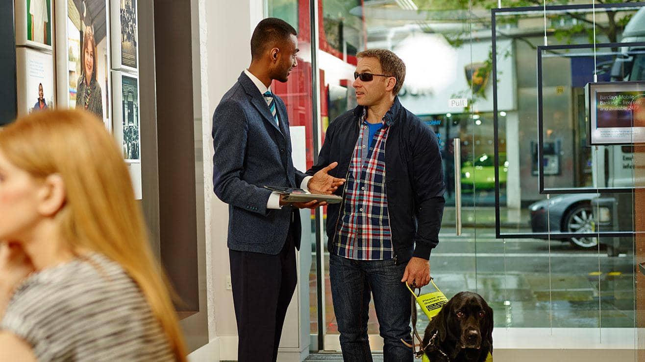 A member of staff speaks to a blind customer who is holding the collar of an assistance dog