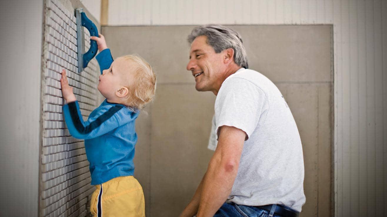 A man lets a child help him to grout some wall tiles 
