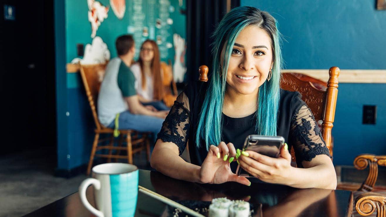 A young woman with blue hair sitting in a café looks up from using her phone