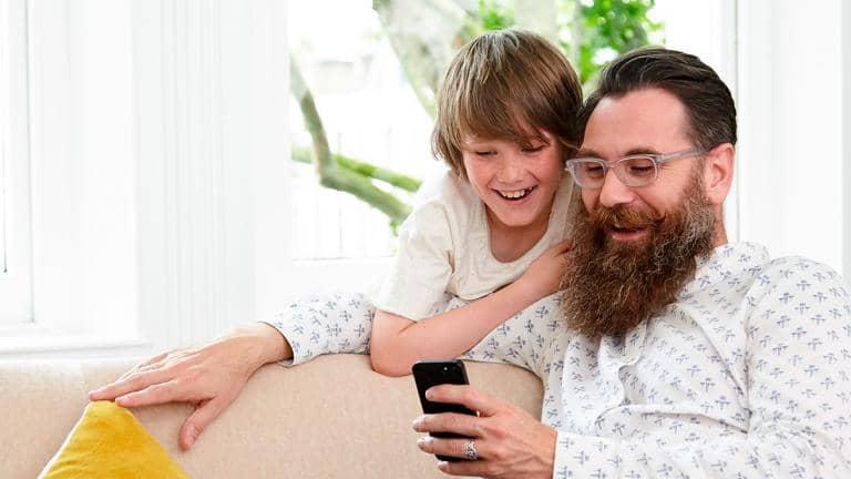 A man sitting on a sofa and a boy standing behind him look at a mobile phone

