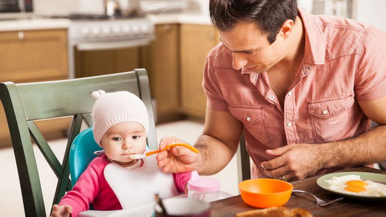 A man feeding a baby in a high chair at a kitchen table