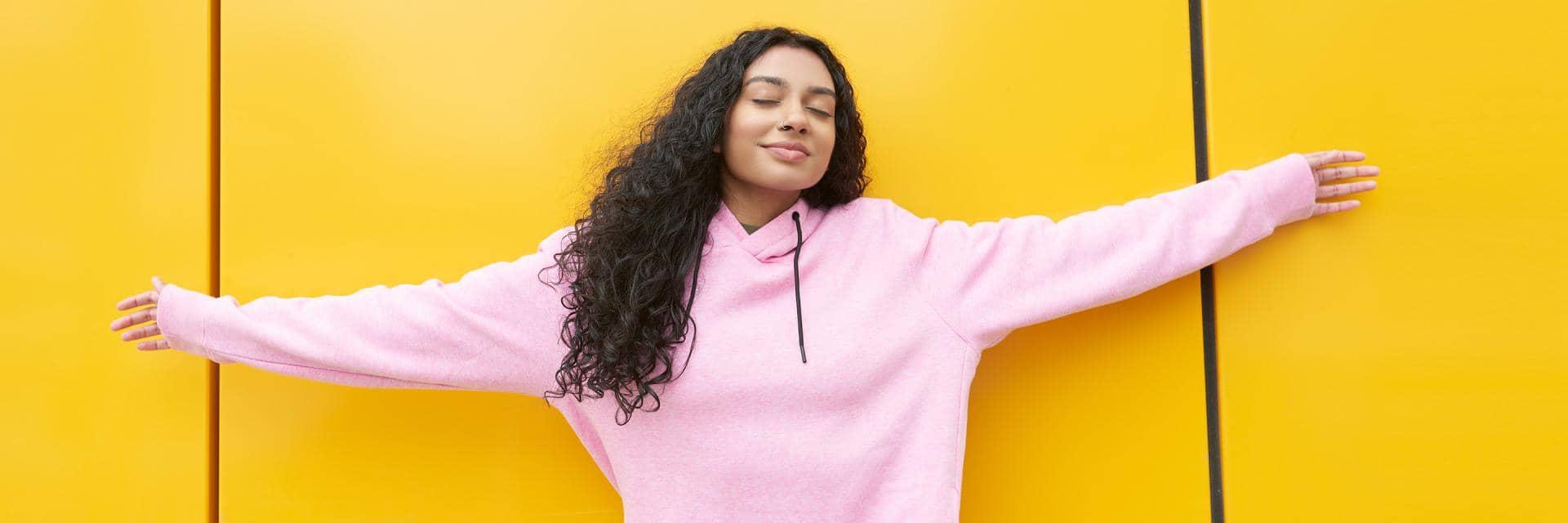Young woman wearing a pink sweatshirt with her arms outstretched in front of a yellow wall