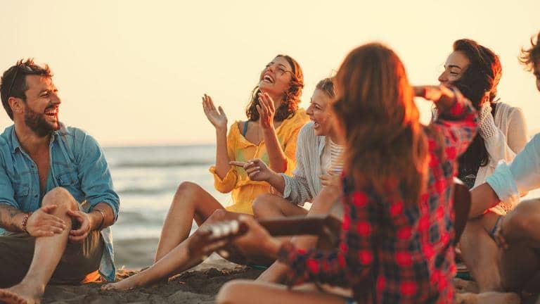 A group of friends sitting on a beach laughing and playing a guitar