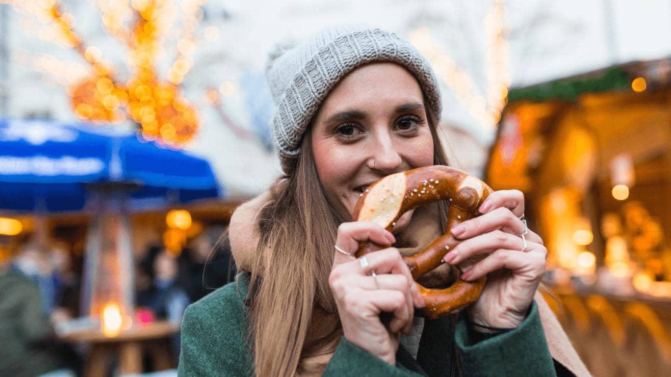 A woman wearing a coat and hat holds up a giant pretzel at a winter market