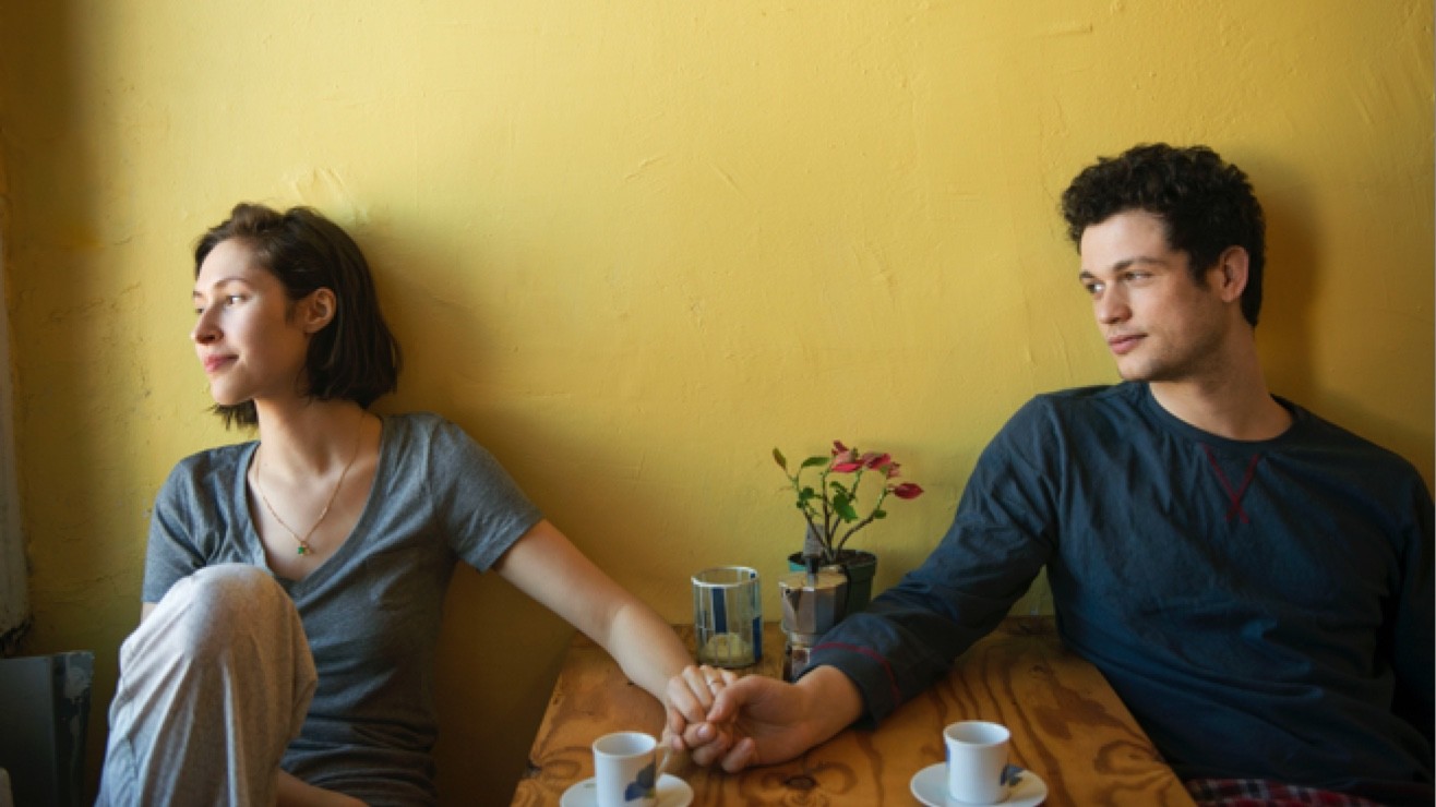 Couple sitting at a kitchen table holding hands and looking off into the distance.